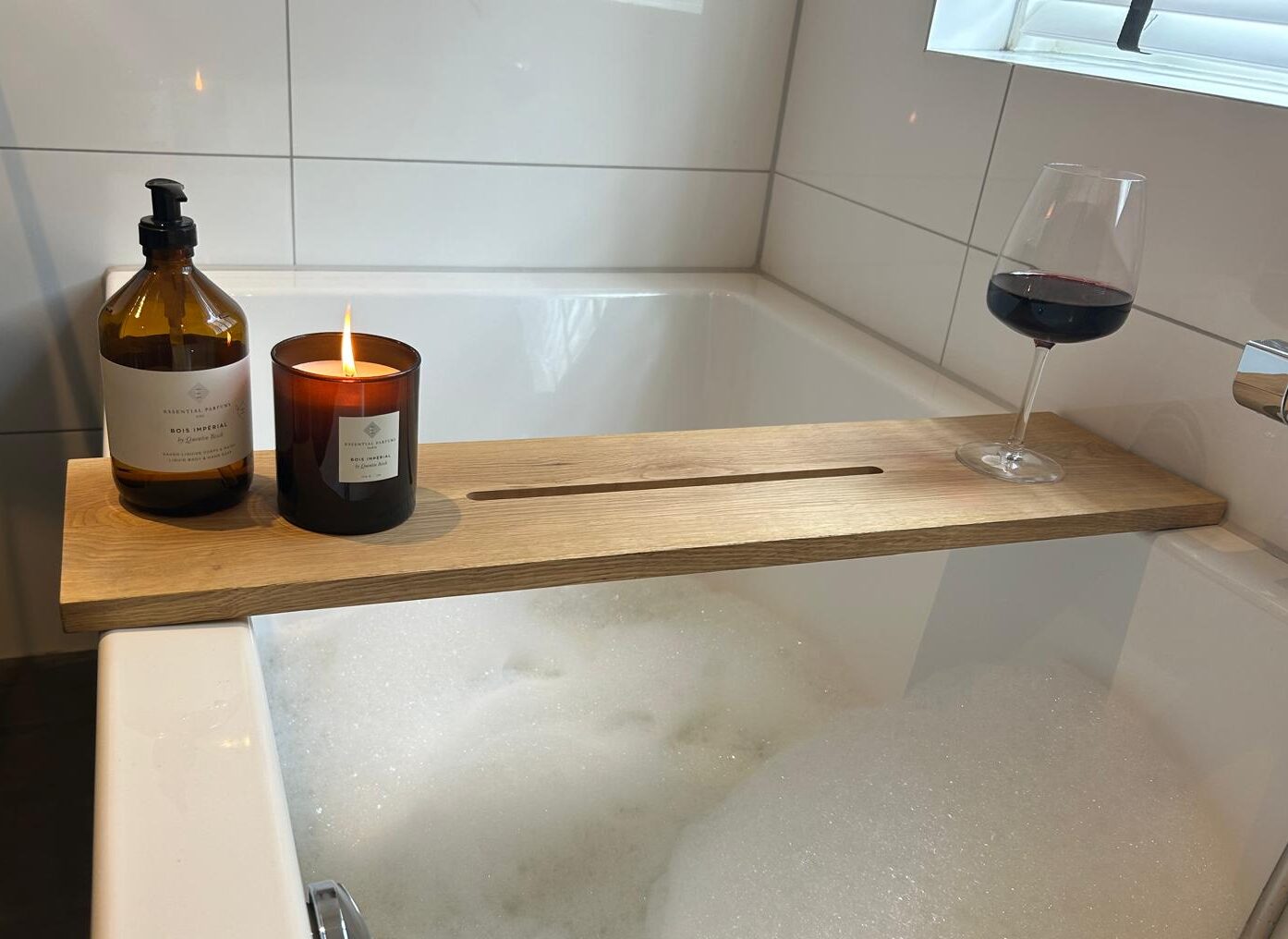 Badplank over een bad met schuim, met een zeeppompje, een brandende kaars en een glas rode wijn