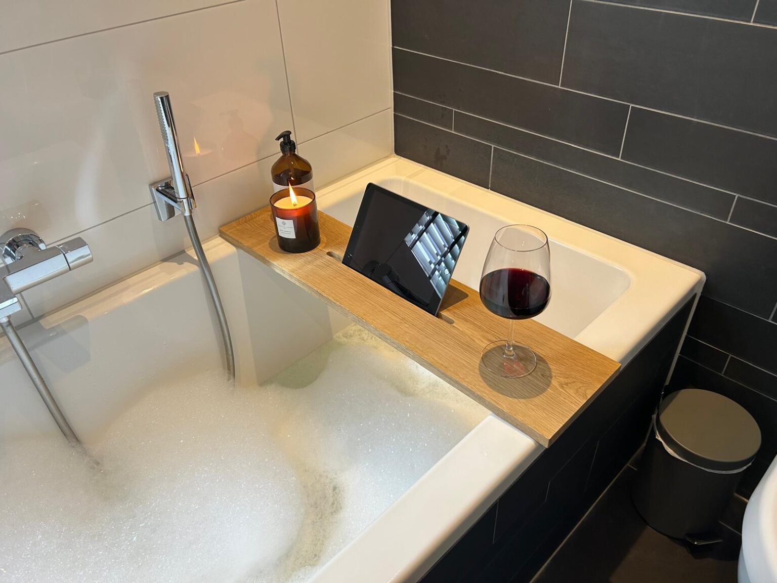 Badplank over een vol bad, met een tablet rechtop in de gleuf, een kaars en een glas rode wijn